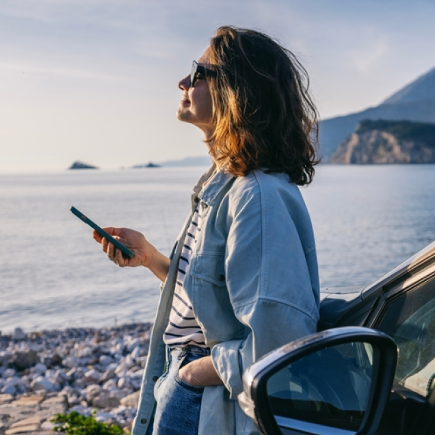 Person standing by a car near a rocky shoreline, holding a smartphone and looking toward the sea.