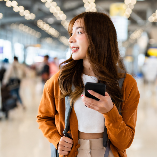 Person in an orange cardigan holding a smartphone in an airport terminal.